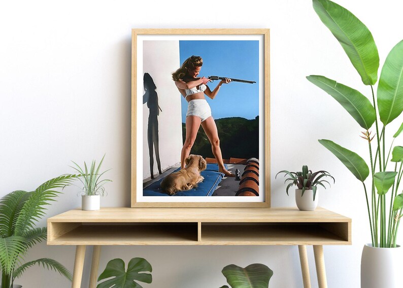 May include: A framed photograph of a woman in a white bikini holding a rifle, standing on a balcony with a small dog lying on a blue cushion.