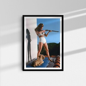 May include: A woman in a white swimsuit holds a rifle and aims it towards the right side of the frame. She is standing on a rooftop with a small dog lying at her feet. The background is a blue sky with some trees in the distance.