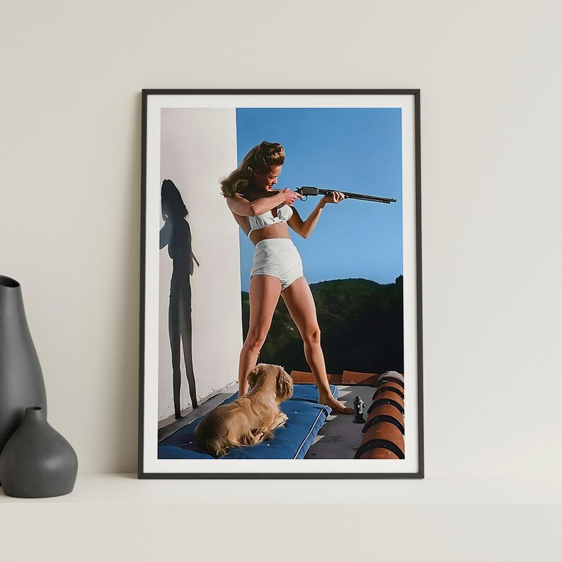 May include: A woman in a white bikini holds a rifle and aims it towards the right side of the frame. She is standing on a patio with a small dog lying on a blue cushion. The background is a blue sky with some trees in the distance.