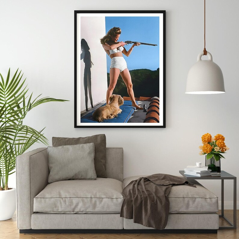 May include: A woman in a white swimsuit holds a rifle and aims it towards the right side of the frame. A small dog sits on a blue cushion behind her. The scene is set against a backdrop of a blue sky and green hills.