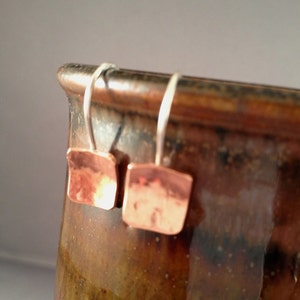 May include: A pair of copper earrings with a hammered finish and a square shape. The earrings are hanging from silver hooks.