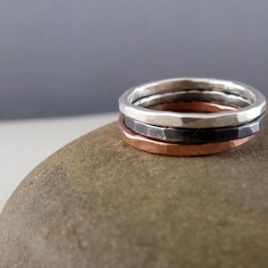 May include: Three hammered metal rings stacked on top of each other. The top ring is silver, the middle ring is black, and the bottom ring is copper.