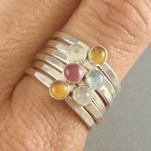 May include: A set of six silver stacking rings with colorful gemstones. The rings feature a variety of colors, including clear, pink, yellow, and blue.