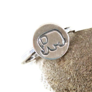 May include: A silver ring with a round, raised elephant design. The ring is on a textured, light brown surface.