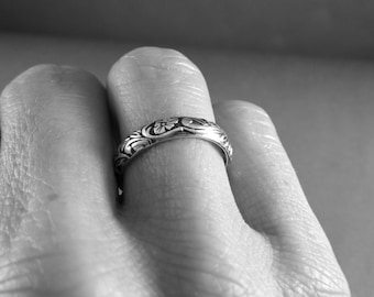 May include: A silver band ring with a floral design. The ring is worn on a person's finger.