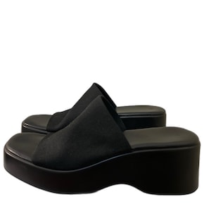 May include: Black platform sandals with a square toe. The sandals have a thick, black sole and a fabric strap across the top of the foot. The shoes are set against a white background.