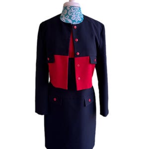 May include: A two-piece women's suit in navy blue and red. The jacket is navy with red accents and buttons. The skirt is navy blue with red pocket details. The suit is displayed on a mannequin.
