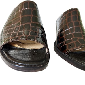 May include: A pair of brown leather slide sandals with a crocodile skin pattern. The sandals have a dark brown sole and a light brown interior. The open-toe design features a strap across the top of the foot.