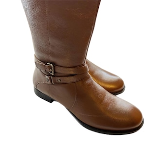 Puede incluir: Un par de botas altas de cuero marr&oacute;n. Las botas tienen una punta redondeada y una correa con hebilla alrededor del tobillo. El cuero tiene un aspecto texturizado y un ligero brillo. Las botas est&aacute;n sobre un fondo blanco.