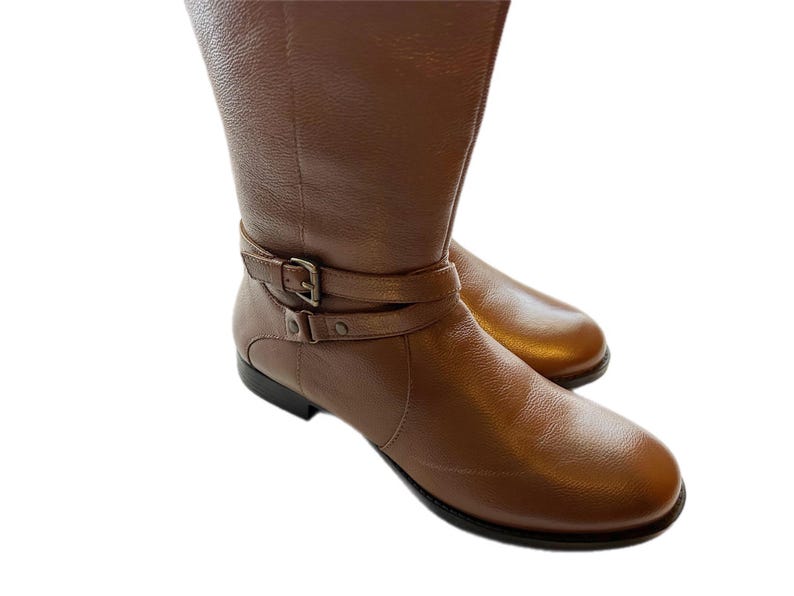 Puede incluir: Un par de botas altas de cuero marr&oacute;n. Las botas tienen una punta redondeada y una correa con hebilla alrededor del tobillo. El cuero tiene un aspecto texturizado y un ligero brillo. Las botas est&aacute;n sobre un fondo blanco.