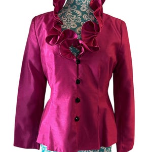 May include: A magenta-colored jacket with a ruffled collar and long sleeves. The jacket has a fitted silhouette and features black buttons down the front. The garment is displayed on a mannequin with a patterned base.