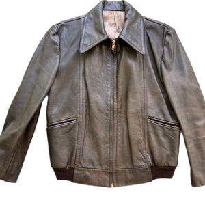 May include: A vintage olive green leather jacket with a zip-up front, two front pockets, and a brown ribbed hem. The jacket has a wide collar and long sleeves. The interior lining is a light tan color.