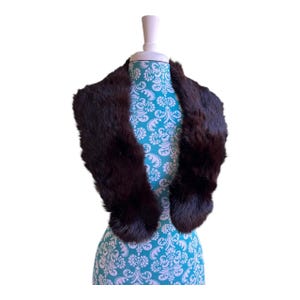 May include: A dark brown fur collar displayed on a mannequin. The collar is a rich, deep brown color and appears to be made of soft fur. The mannequin is covered in a teal fabric with a white floral pattern.