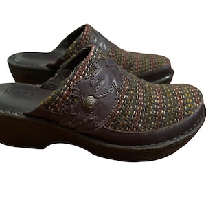 May include: A pair of brown leather clogs featuring a multi-colored woven fabric upper with a dark brown leather overlay. The overlay is decorated with a floral-shaped cutout and a small metallic stud. The clogs have a dark brown sole.