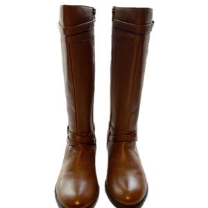 Puede incluir: Un par de botas altas de cuero marr&oacute;n. Las botas tienen una punta redondeada, un tac&oacute;n peque&ntilde;o y correas decorativas alrededor del tobillo y la pantorrilla. Cada bota tiene una cremallera lateral. El cuero tiene un ligero brillo.
