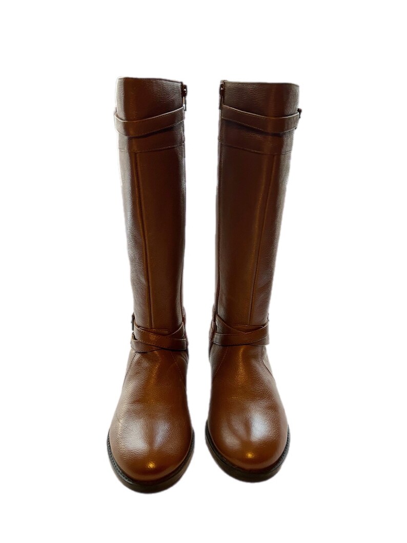 Puede incluir: Un par de botas altas de cuero marr&oacute;n. Las botas tienen una punta redondeada, un tac&oacute;n peque&ntilde;o y correas decorativas alrededor del tobillo y la pantorrilla. Cada bota tiene una cremallera lateral. El cuero tiene un ligero brillo.