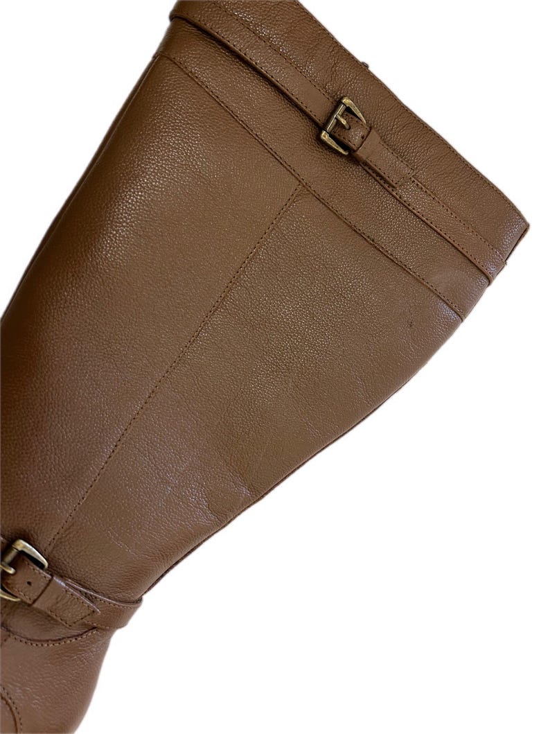 Puede incluir: Primer plano de una bota de montar de cuero marr&oacute;n. La bota presenta una superficie texturizada, una correa con una hebilla dorada en la parte superior y otra hebilla en el tobillo. La bota est&aacute; en &aacute;ngulo, mostrando su dise&ntilde;o.