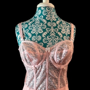 May include: A light pink lace bustier with adjustable straps and a small bow detail. The bustier is displayed on a mannequin with a teal and white floral pattern. The garment has a structured design with a satin-like trim.