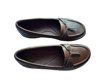 May include: A pair of brown leather loafers featuring a rounded toe, a decorative tassel detail on the vamp, and a low heel. The interior footbed is dark brown and includes the visible brand name Clarks printed in a light color on the heel area.