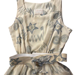 May include: A sleeveless, cream-colored dress with a blue and white abstract print. The dress has a white belt with a decorative buckle and a blue bow.