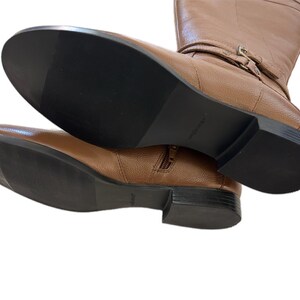 Puede incluir: Un par de botas altas de cuero marr&oacute;n. Las botas tienen una cremallera lateral, un detalle de hebilla y una suela negra. Las botas se muestran desde abajo, destacando las suelas y el cuero texturizado.