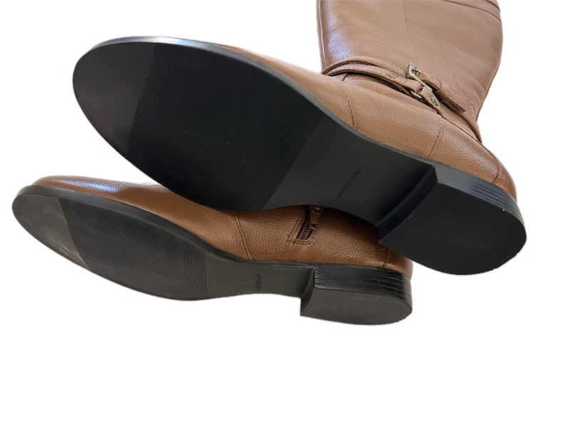 Puede incluir: Un par de botas altas de cuero marr&oacute;n. Las botas tienen una cremallera lateral, un detalle de hebilla y una suela negra. Las botas se muestran desde abajo, destacando las suelas y el cuero texturizado.
