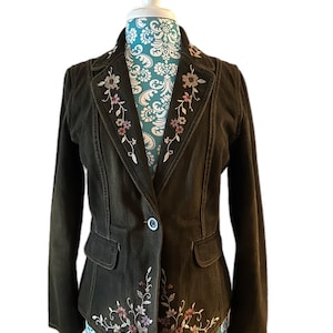 May include: A dark olive green blazer with floral embroidery. The blazer features a single button closure, two front pockets, and embroidered floral designs along the lapels and front. The jacket is displayed on a mannequin.