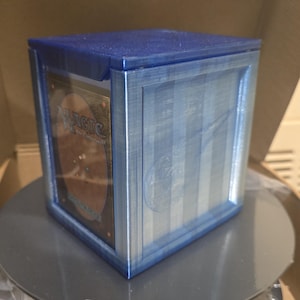 May include: A blue, 3D-printed card storage box. The box has a square shape with a lid and is designed to hold trading cards. The front of the box has a vertical striped design. The box is sitting on a gray surface.