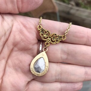 May include: Gold tone necklace with a teardrop shaped pendant featuring a clear faceted stone. The pendant is attached to a delicate chain with a filigree design.