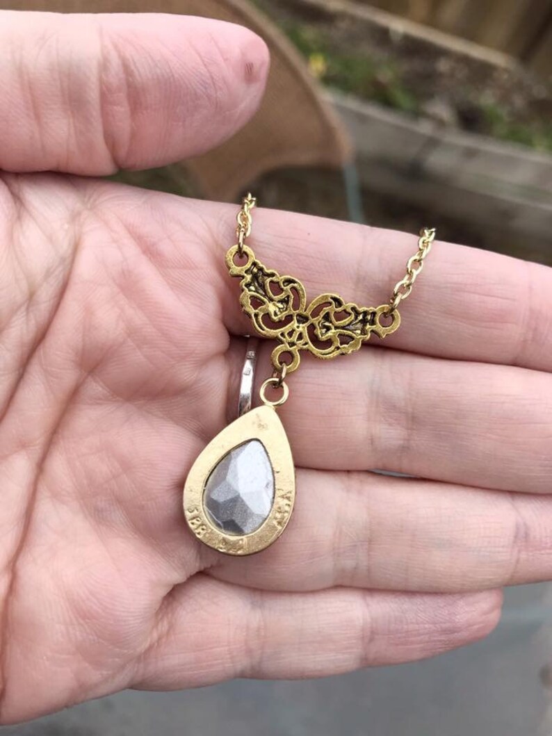 May include: Gold tone necklace with a teardrop shaped pendant featuring a clear faceted stone. The pendant is attached to a delicate chain with a filigree design.