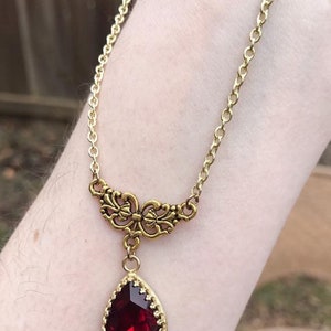 May include: A gold chain necklace with a teardrop-shaped red gemstone pendant. The pendant is surrounded by a gold filigree design.