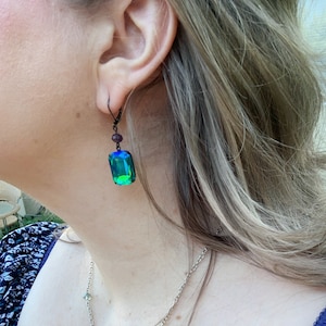 Puede incluir: Primer plano de una persona que lleva un pendiente de metal oscuro. El pendiente presenta un gran cristal rectangular, iridiscente verde y azul. Una cuenta p&uacute;rpura m&aacute;s peque&ntilde;a est&aacute; encima del cristal. El pendiente est&aacute; sujeto a un gancho de metal oscuro.