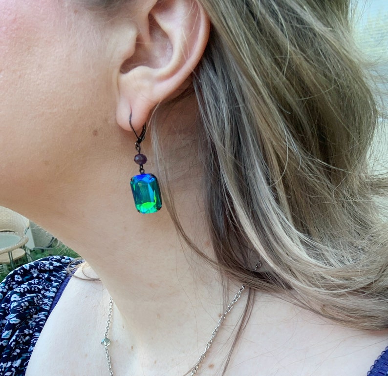 Puede incluir: Primer plano de una persona que lleva un pendiente de metal oscuro. El pendiente presenta un gran cristal rectangular, iridiscente verde y azul. Una cuenta p&uacute;rpura m&aacute;s peque&ntilde;a est&aacute; encima del cristal. El pendiente est&aacute; sujeto a un gancho de metal oscuro.