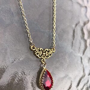 May include: A gold chain necklace with a red teardrop-shaped gemstone pendant. The pendant is surrounded by a gold filigree design.