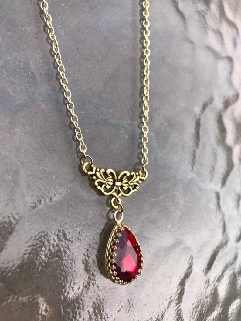May include: A gold chain necklace with a red teardrop-shaped gemstone pendant. The pendant is surrounded by a gold filigree design.
