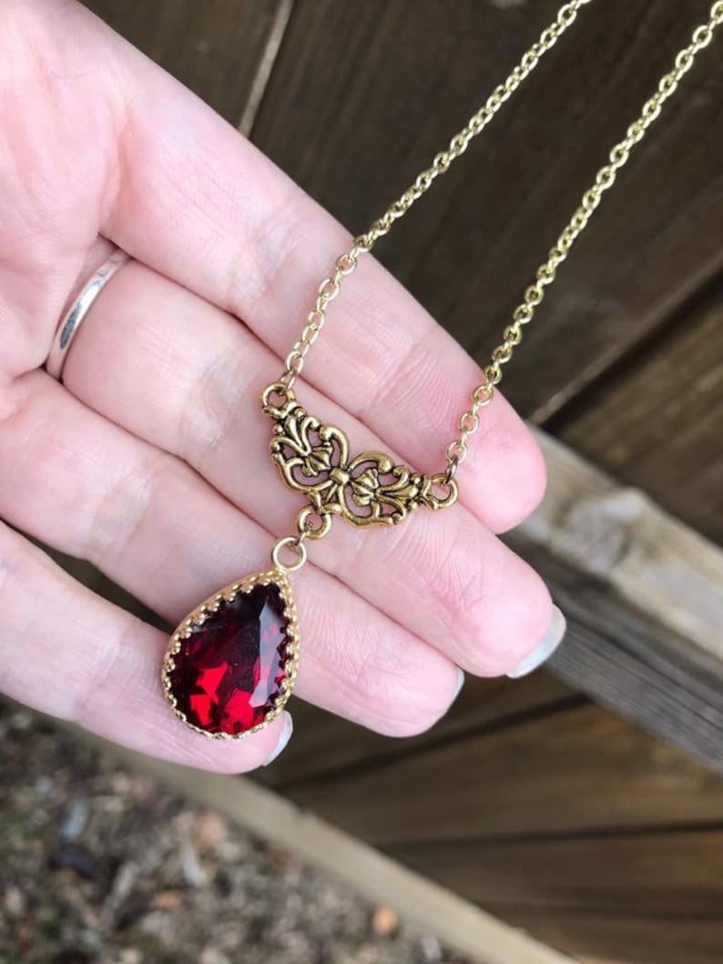 May include: A gold chain necklace with a red teardrop-shaped gemstone pendant. The pendant is surrounded by a gold filigree design.