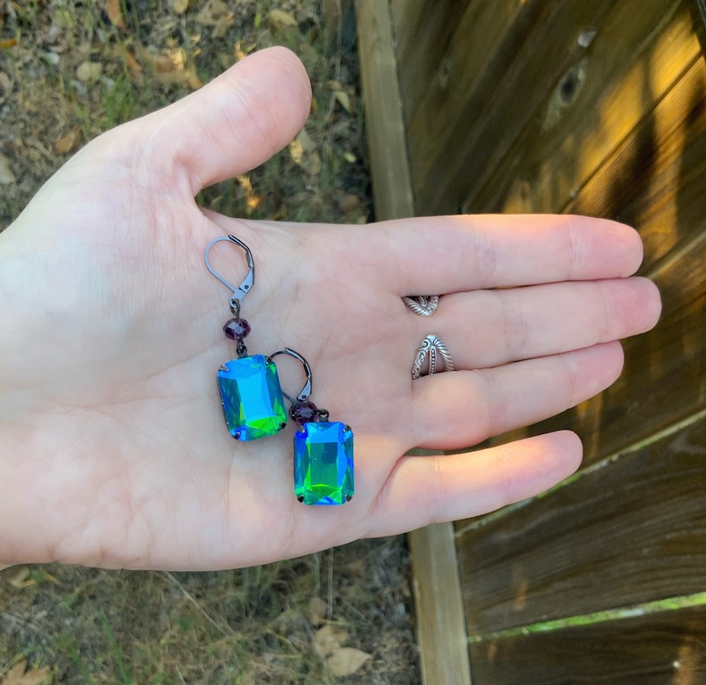 Puede incluir: Un par de pendientes con cierre de palanca y cristales iridiscentes rectangulares con talla esmeralda. Los cristales cambian de azul a verde. Peque&ntilde;as cuentas moradas acent&uacute;an la parte superior de cada pendiente. El metal es de color plomo oscuro.