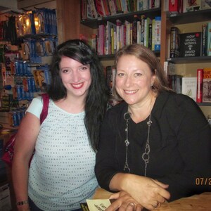 Puede incluir: Dos mujeres sonriendo en una librer&iacute;a. Una tiene el pelo largo y oscuro y lleva una blusa azul claro. La otra tiene el pelo casta&ntilde;o claro y lleva una blusa negra y un collar plateado. Estanter&iacute;as llenas de libros son visibles en el fondo.