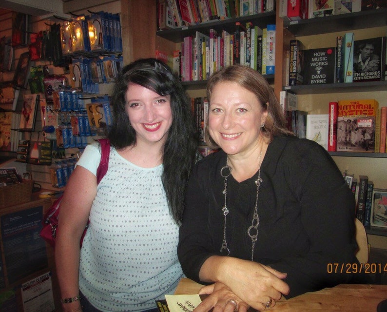 Puede incluir: Dos mujeres sonriendo en una librer&iacute;a. Una tiene el pelo largo y oscuro y lleva una blusa azul claro. La otra tiene el pelo casta&ntilde;o claro y lleva una blusa negra y un collar plateado. Estanter&iacute;as llenas de libros son visibles en el fondo.