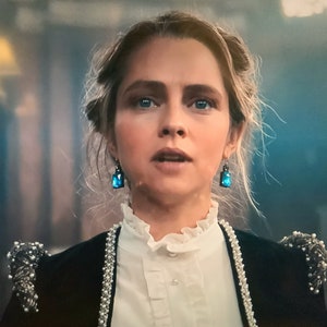 Puede incluir: Una mujer con pendientes de piedras preciosas azules y una chaqueta negra con adornos de perlas. La chaqueta tiene ribete de perlas y detalles decorativos en los hombros. Lleva una blusa blanca con cuello con volantes.