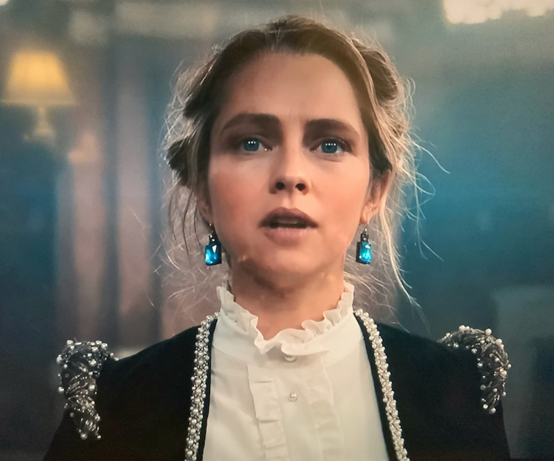 Puede incluir: Una mujer con pendientes de piedras preciosas azules y una chaqueta negra con adornos de perlas. La chaqueta tiene ribete de perlas y detalles decorativos en los hombros. Lleva una blusa blanca con cuello con volantes.