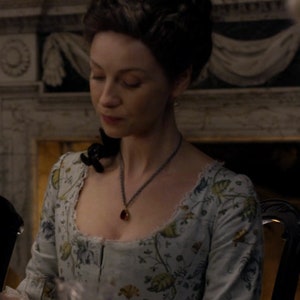 May include: A woman wearing a light blue dress with a floral pattern. She is wearing a necklace with a red gemstone pendant. The dress has a low neckline and a fitted bodice.