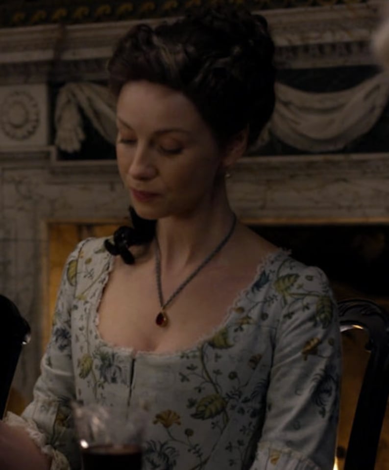 May include: A woman wearing a light blue dress with a floral pattern. She is wearing a necklace with a red gemstone pendant. The dress has a low neckline and a fitted bodice.