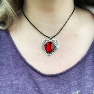 Könnte beinhalten: Eine silberne Halskette mit einem herzförmigen Anhänger, der mit einem großen roten Edelstein besetzt ist. Die Kette hat eine schwarze Schnur.