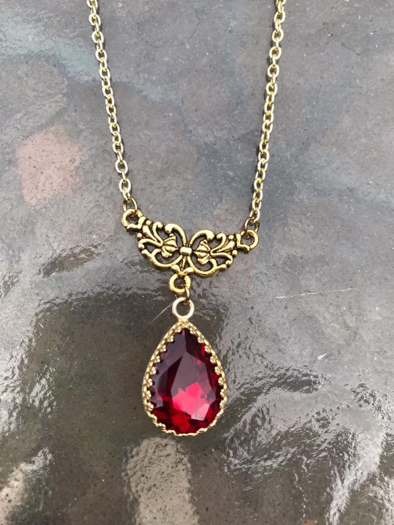 May include: A gold chain necklace with a red teardrop-shaped gemstone pendant. The pendant is surrounded by a gold filigree design.