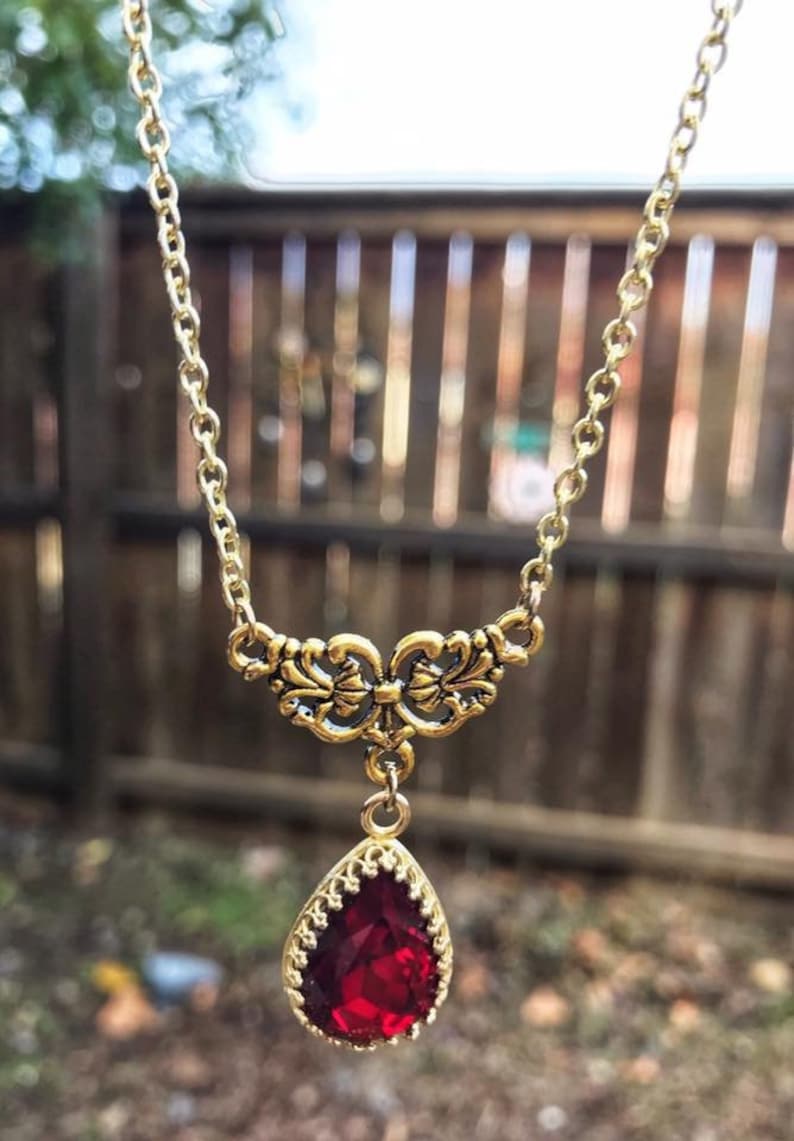 May include: A gold chain necklace with a red teardrop-shaped gemstone pendant. The pendant is surrounded by a gold filigree design.