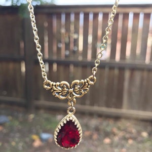May include: A gold chain necklace with a red teardrop-shaped pendant. The pendant is set in a gold filigree design.