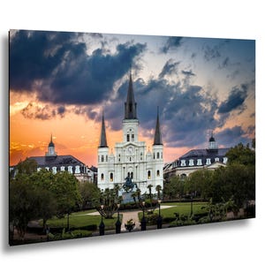 Puede incluir: Fotografía de una catedral blanca con altas agujas, un reloj y una estatua en frente. El cielo está lleno de nubes dramáticas y una colorida puesta de sol. Árboles verdes y céspedes cuidados rodean el edificio.
