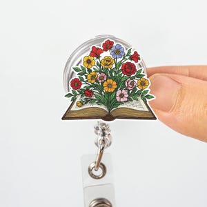 Puede incluir: Un carrete de insignia con un diseño floral. La imagen presenta un colorido ramo de flores que emerge de un libro abierto. Las flores son rojas, amarillas, azules y rosas. El carrete tiene una carcasa de plástico transparente y un clip de metal.