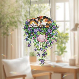 May include: A decorative hanging cat ornament. The cat is sleeping on purple flowers in a pink planter. The cat has orange, black, and white fur. The ornament hangs from a silver chain. The background is a blurred image of a room.